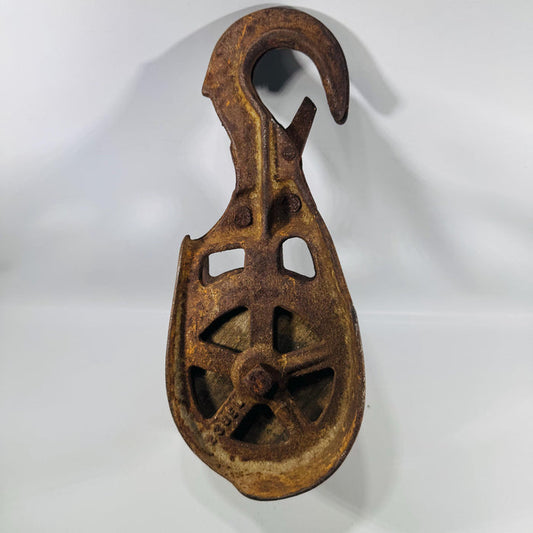 Vintage cast iron single-wheel barn pulley with large fixed hook and safety latch, viewed from the front, showing open spoked iron wheel frame, deep rust and ochre patina throughout, a